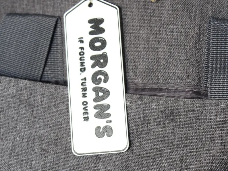 A white Kayem.co backpack return tag reading "Morgan’s, if found turn over" clipped to a grey backpack