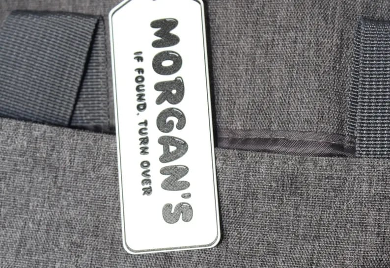 A white Kayem.co backpack return tag reading Morgan’s, if found turn over, clipped to a grey backpack