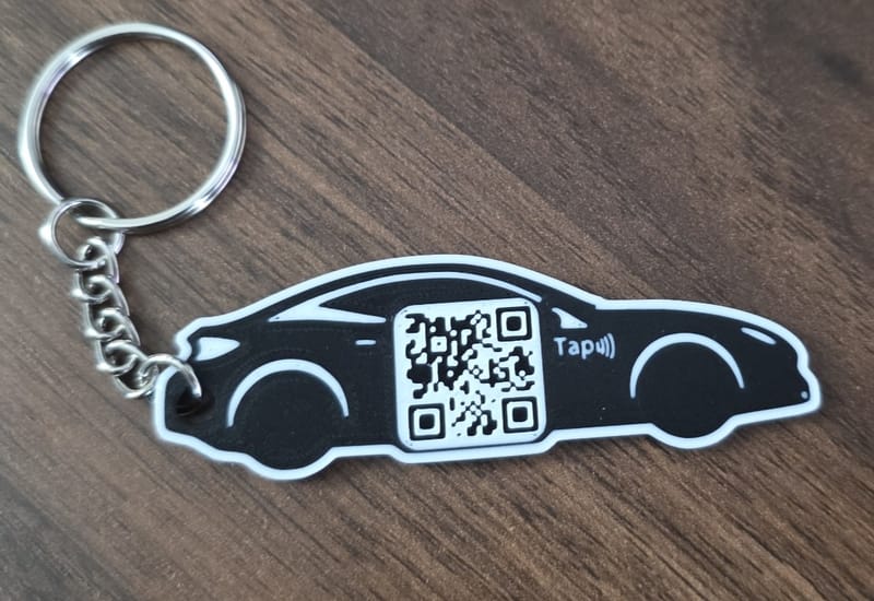Black car-shaped Kayem.co keychain with a QR code and tap symbol on a wood table