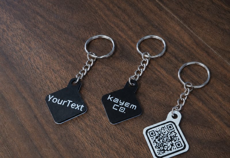 Three handmade Kayem.co keychains on a wood table, showing custom text, logo, and QR code tags